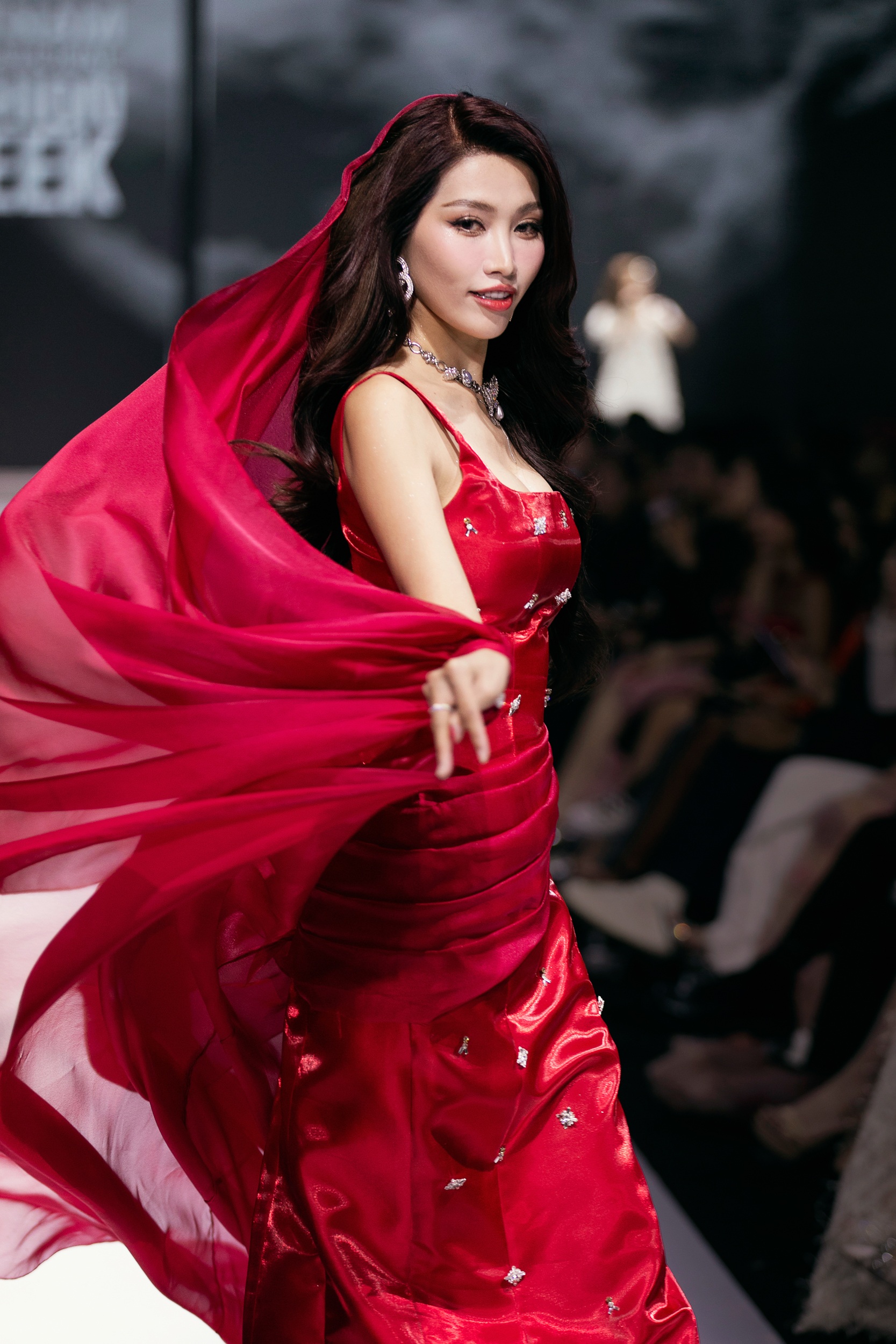 Quỳnh Châu khoác bộ đầm đỏ ôm sát được "đo ni đóng giày" theo số đo, tôn trọn vẹn vóc dáng. Trang phục dùng chất liệu tafta, đính kết điểm xuyết đá lấp lánh, phối cùng áo choàng tạo vẻ quyền lực như nữ thần. 
