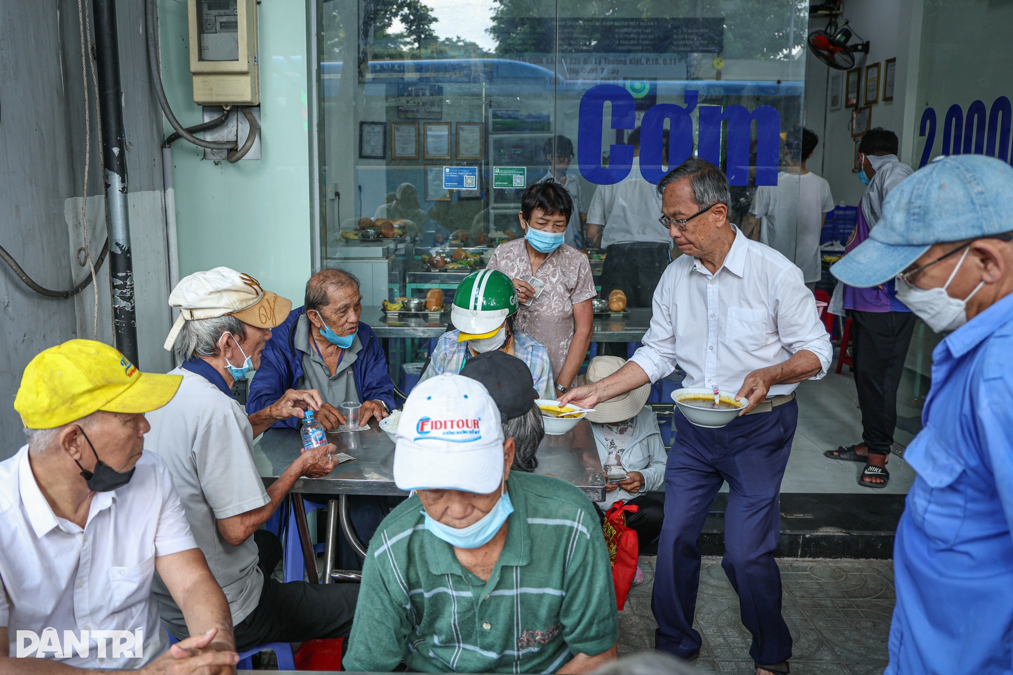 Vào mỗi trưa, nắng như đổ lửa xuống mặt đường Lý Thường Kiệt, trước một quán cơm xã hội nhỏ không biển hiệu, hàng dài người vẫn lặng lẽ xếp hàng. Từ người già, người khuyết tật, công nhân, sinh viên, lao động tự do... Họ đứng kín một khoảng vỉa hè, tay che nắng, mắt hướng vào bên trong căn bếp nhỏ chờ đợi một bữa ăn đủ đầy chỉ với giá 2.000 đồng. Giữa dòng người chen chúc ấy, xuất hiện bóng dáng của một ông lão tóc bạc trắng, mặc áo sơ mi trắng chỉnh tề. Ông chậm rãi bưng từng khay cơm đến tận bàn, cúi nhẹ đầu cảm ơn rồi quay lại vào gian bếp, tiếp tục sắp xếp đồ ăn lên khay.